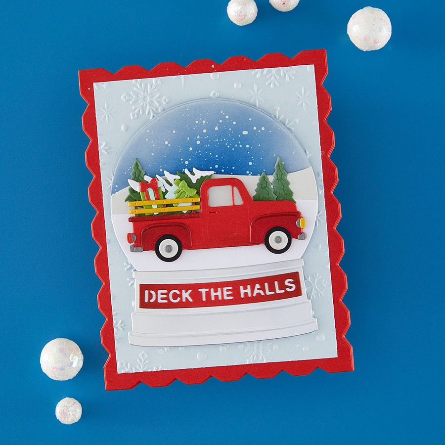 A handmade Christmas card featuring a red pickup truck carrying Christmas trees inside a clear snow globe design, with the text 'DECK THE HALLS' on a white banner below. The card has a scalloped red border and is displayed on a bright blue background with decorative white paper spheres.