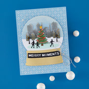 A handmade square greeting card featuring a die-cut snow globe design with a Christmas tree, city skyline, and silhouettes of people walking. The card has a light blue patterned border and a gold banner reading 'MERRY MOMENTS' on a bright blue background, with small white pom-poms scattered around.