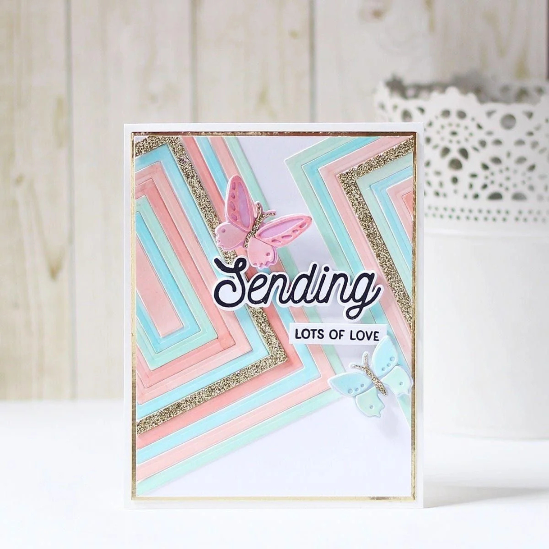 A handmade greeting card with layered pastel pink, light blue, and white geometric stripes, glitter accents, and two butterfly embellishments. The card is displayed upright on a white surface against a light wood panel background. Using Spellbinders Precision Layering A2 A Etched Dies