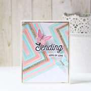 A handmade greeting card with layered pastel pink, light blue, and white geometric stripes, glitter accents, and two butterfly embellishments. The card is displayed upright on a white surface against a light wood panel background. Using Spellbinders Precision Layering A2 A Etched Dies