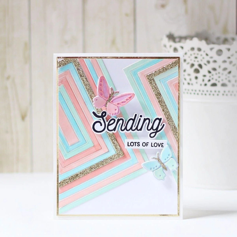 A handmade greeting card with layered pastel pink, light blue, and white geometric stripes, glitter accents, and two butterfly embellishments. The card is displayed upright on a white surface against a light wood panel background. Using Spellbinders Precision Layering A2 A Etched Dies