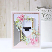A handmade greeting card with a floral design, featuring pink and mint green paper flowers, a gold inner frame, and the words 'SENDING HUGS' on a black banner, set against a soft, light background.