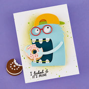 A handmade greeting card featuring a blue monster character wearing red glasses and a yellow cap, holding a pink frosted donut. The card has green paint splatters, the text 'I licked it IT'S MINE' at the bottom, and is displayed on a purple background next to a cookie cutout.