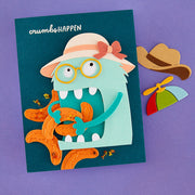 A handmade layered paper card featuring a cartoon monster wearing a straw hat and yellow glasses, surrounded by orange bananas and small yellow bugs, displayed on a purple background with matching paper accessories.