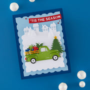 A handmade layered paper holiday card featuring a green pickup truck carrying Christmas trees and gifts, set against a snowy winter scene with houses and a red banner reading 'TIS THE SEASON'. The card is displayed on a blue background with decorative white pom-poms.