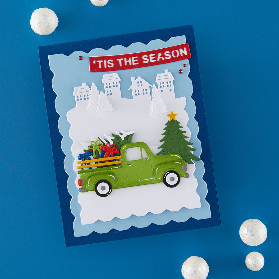 A handmade layered paper holiday card featuring a green pickup truck carrying Christmas trees and gifts, set against a snowy winter scene with houses and a red banner reading 'TIS THE SEASON'. The card is displayed on a blue background with decorative white pom-poms.