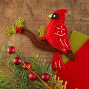 A red felt cardinal bird decoration perched on a brown felt branch, decorated with white stitched details, green felt holly leaves with red berries, and placed on a wooden surface with additional greenery and red berries.