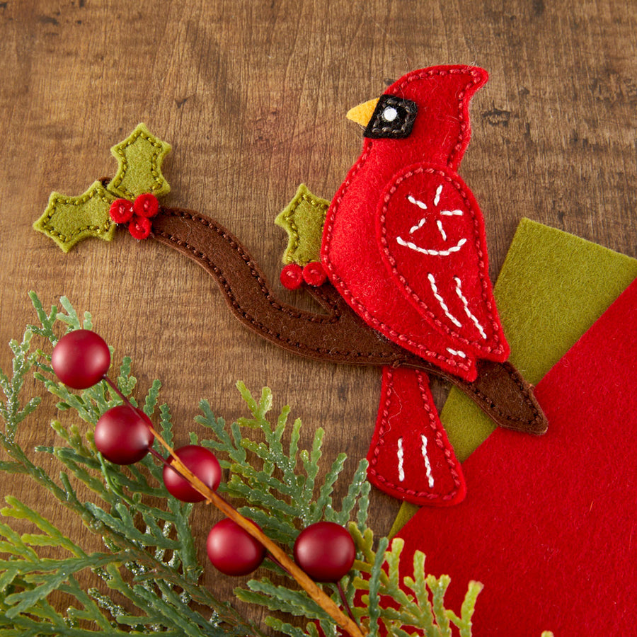 A red felt cardinal bird decoration perched on a brown felt branch, decorated with white stitched details, green felt holly leaves with red berries, and placed on a wooden surface with additional greenery and red berries.
