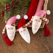Three handmade felt Christmas gnome ornaments with red and pink hats, white beards, and red tassels, arranged on a rustic wooden surface next to a wicker basket and red ribbons.