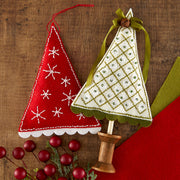 Two handmade felt Christmas tree felt ornaments on wooden sticks, displayed on a rustic wood surface with red berries and a red fabric background. One ornament is red with white embroidered snowflakes and a red hanging loop.