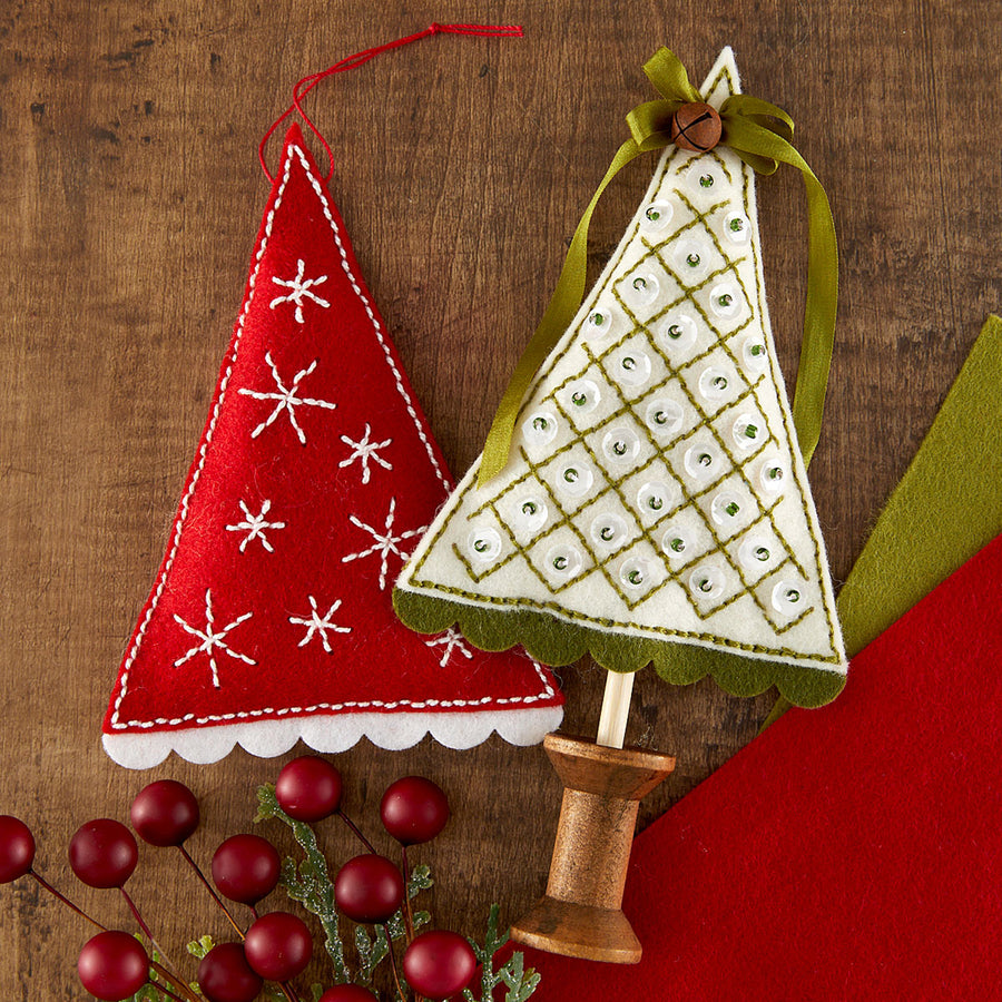 Two handmade felt Christmas tree felt ornaments on wooden sticks, displayed on a rustic wood surface with red berries and a red fabric background. One ornament is red with white embroidered snowflakes and a red hanging loop.