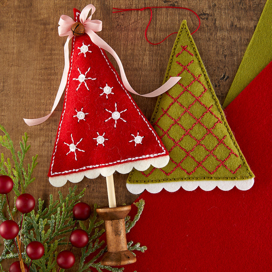 Two handmade felt Christmas tree ornaments on wooden sticks, styled with evergreen branches and red berries on a rustic wood background. One ornament is red with white embroidered snowflakes and a pink ribbon topper, and the other is olive green with a red diamond pattern and white scalloped edge.