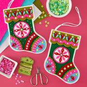 Two colorful handmade felt Christmas stockings with embroidered patterns, shown on a bright pink background with craft supplies including sequins, scissors, thread, and ribbon.