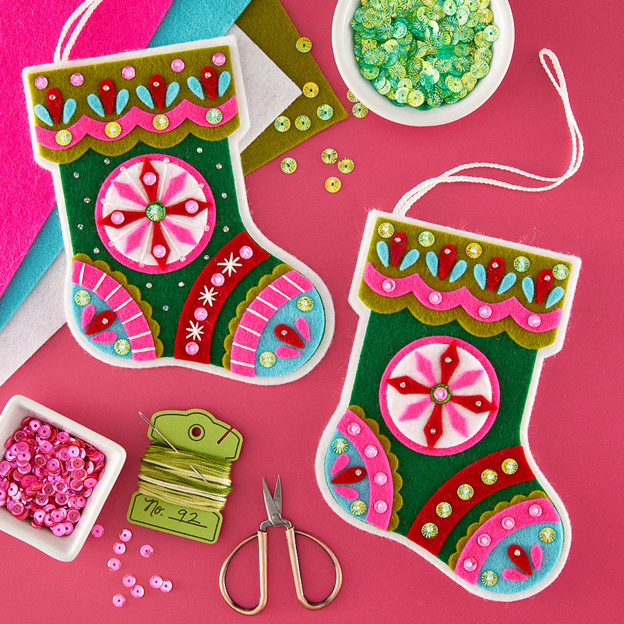 Two colorful handmade felt Christmas stockings with embroidered patterns, shown on a bright pink background with craft supplies including sequins, scissors, thread, and ribbon.