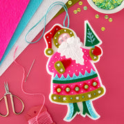 A colorful handmade felt Christmas ornament shaped like Santa Claus, shown on a bright pink background with craft supplies including scissors, thread, felt sheets, and sequins.