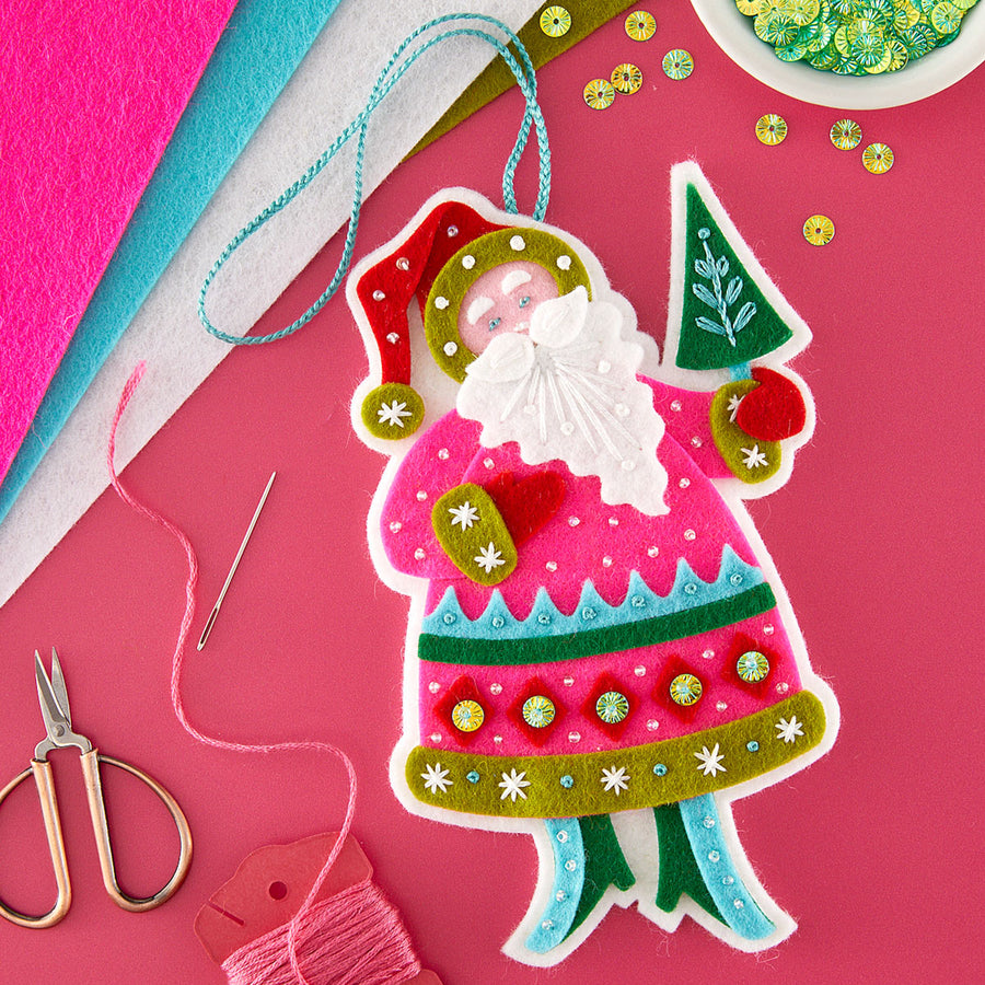 A colorful handmade felt Christmas ornament shaped like Santa Claus, shown on a bright pink background with craft supplies including scissors, thread, felt sheets, and sequins.