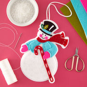 A handmade felt snowman ornament laid flat on a bright pink background with craft supplies around it, including thread, scissors, a bowl of white beads, and colored felt sheets.