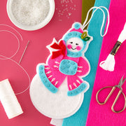 A festive handmade snowman ornament made from felt and fabric, displayed on a bright pink background with craft supplies including thread, scissors, beads, and colored felt sheets.