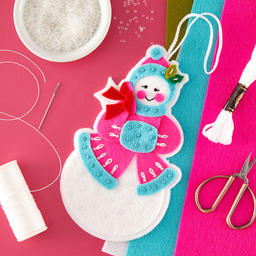 A festive handmade snowman ornament made from felt and fabric, displayed on a bright pink background with craft supplies including thread, scissors, beads, and colored felt sheets.