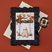 A handmade greeting card with an ornate embossed black frame holding a Halloween card front with a ghost on a swing and the words 'STAY SPOOKY, FRIEND,' styled on a red background with cream envelopes and a small matching card holder.