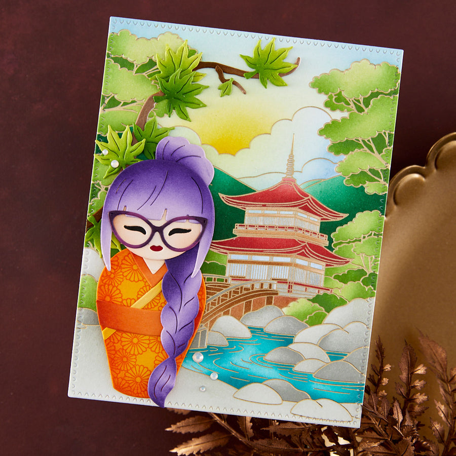 A rectangular handmade card featuring a stylized Japanese scene with a purple-haired doll in an orange kimono, a pagoda, trees, a bridge, and a blue river, photographed against a dark brown background with decorative dried leaves and a gold-toned object at the right edge.