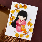 A white rectangular card featuring a stylized paper-cut illustration of a girl with black hair in a pink kimono, holding an orange lantern, surrounded by yellow maple leaves and small gold dots, displayed on a dark brown surface with decorative dried foliage.
