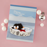 A handmade Christmas greeting card featuring a 3D layered penguin on an iceberg, styled on a pink background with decorative white and silver ornaments.