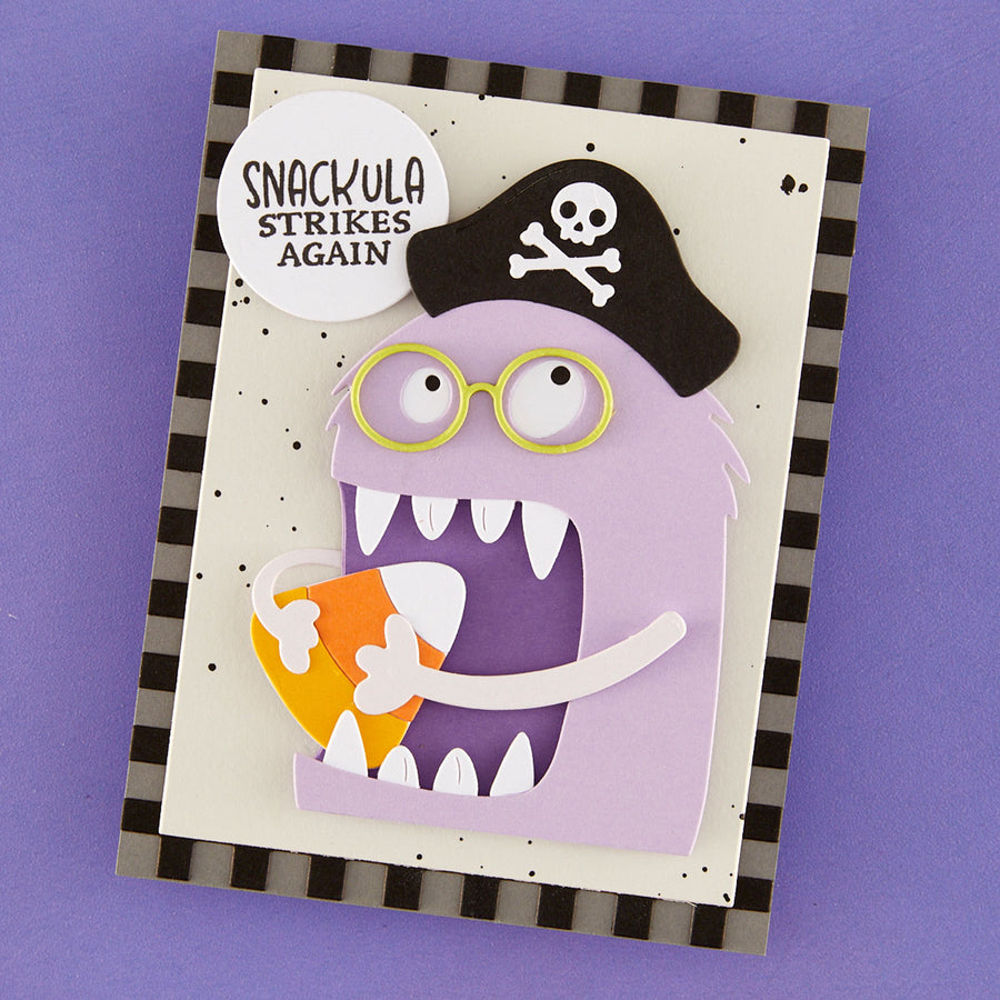 A handmade layered paper card featuring a purple monster wearing a black pirate hat with a skull and crossbones, yellow-rimmed glasses, and holding a candy corn. A white circular badge reads 'SNACKULA STRIKES AGAIN'.