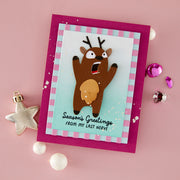 A handmade Christmas greeting card featuring a cartoon reindeer with its mouth wide open, surrounded by a pink and light blue striped border and a magenta card base. The card is displayed on a pink background with small decorative ornaments.