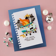 A handmade layered paper craft card featuring a raccoon and cat on a light blue boat with colorful Christmas lights and flames, set on a blue cardstock background with the text 'THE LIGHTS REALLY THE IT TOGETHER'. The card is styled on a pink background with holiday ornaments.