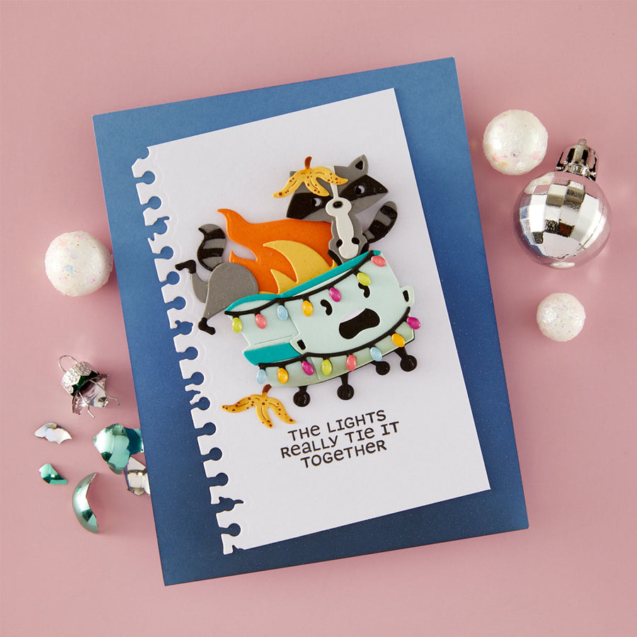 A handmade layered paper craft card featuring a raccoon and cat on a light blue boat with colorful Christmas lights and flames, set on a blue cardstock background with the text 'THE LIGHTS REALLY THE IT TOGETHER'. The card is styled on a pink background with holiday ornaments.