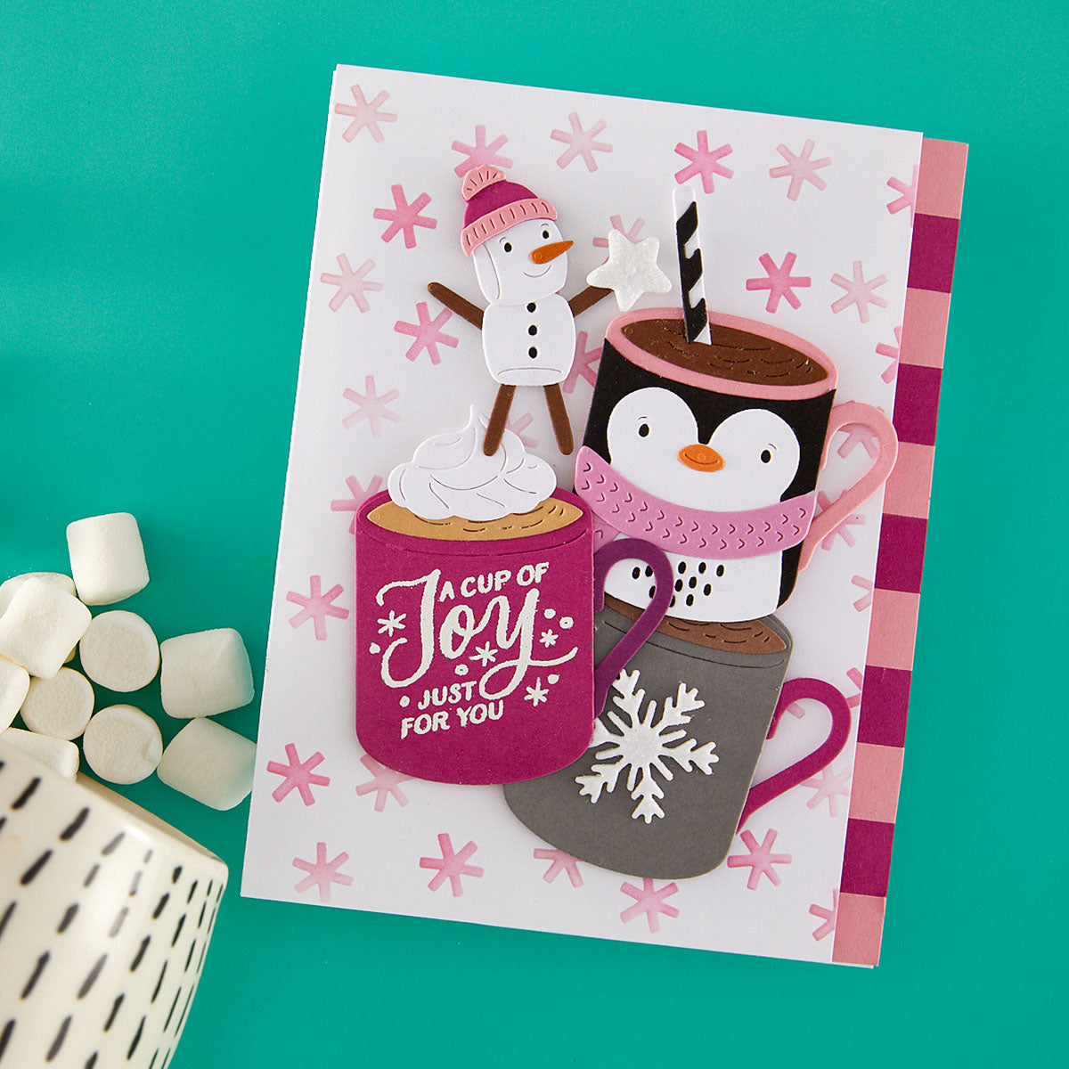 A handmade holiday greeting card featuring cartoon penguins and a snowman with hot cocoa cups, styled on a teal background with marshmallows and a ceramic mug from Spellbinders using the Mug Friends Etched Dies and the Spellbinders Snow Joy Stencil
