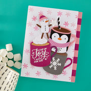 A handmade holiday greeting card featuring cartoon penguins and a snowman with hot cocoa cups, styled on a teal background with marshmallows and a ceramic mug from Spellbinders using the Mug Friends Etched Dies and the Spellbinders Snow Joy Stencil