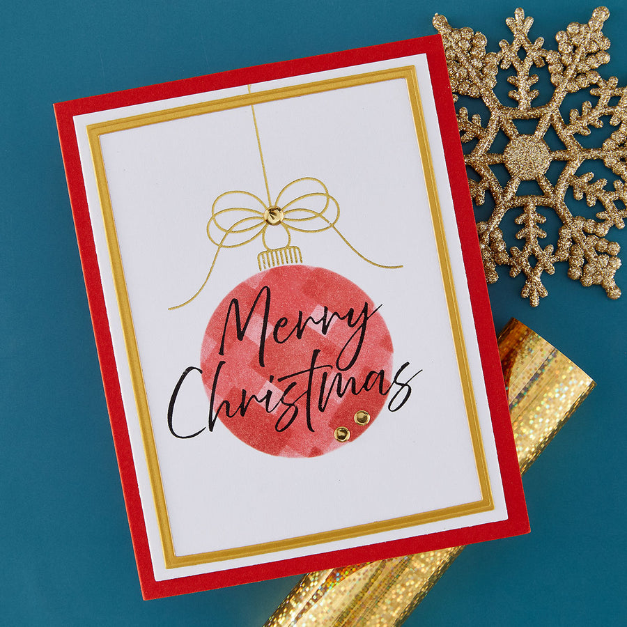 A handmade festive Christmas greeting card with a red border, gold inner border, and a red watercolor ornament illustration with the words 'Merry Christmas' in black script. The card is photographed on a teal background with gold decorative snowflake and ribbon props.