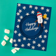 A handmade navy blue handmade holiday card with a circular cutout in the center and the words 'Happy Holidays' in white script. The card is decorated with white snowflakes and features a paper snowman wearing a red and green hat and brown arms, holding a yellow star. Spellbinders Mugs and Kisses Sentiments Clear Stamp Set