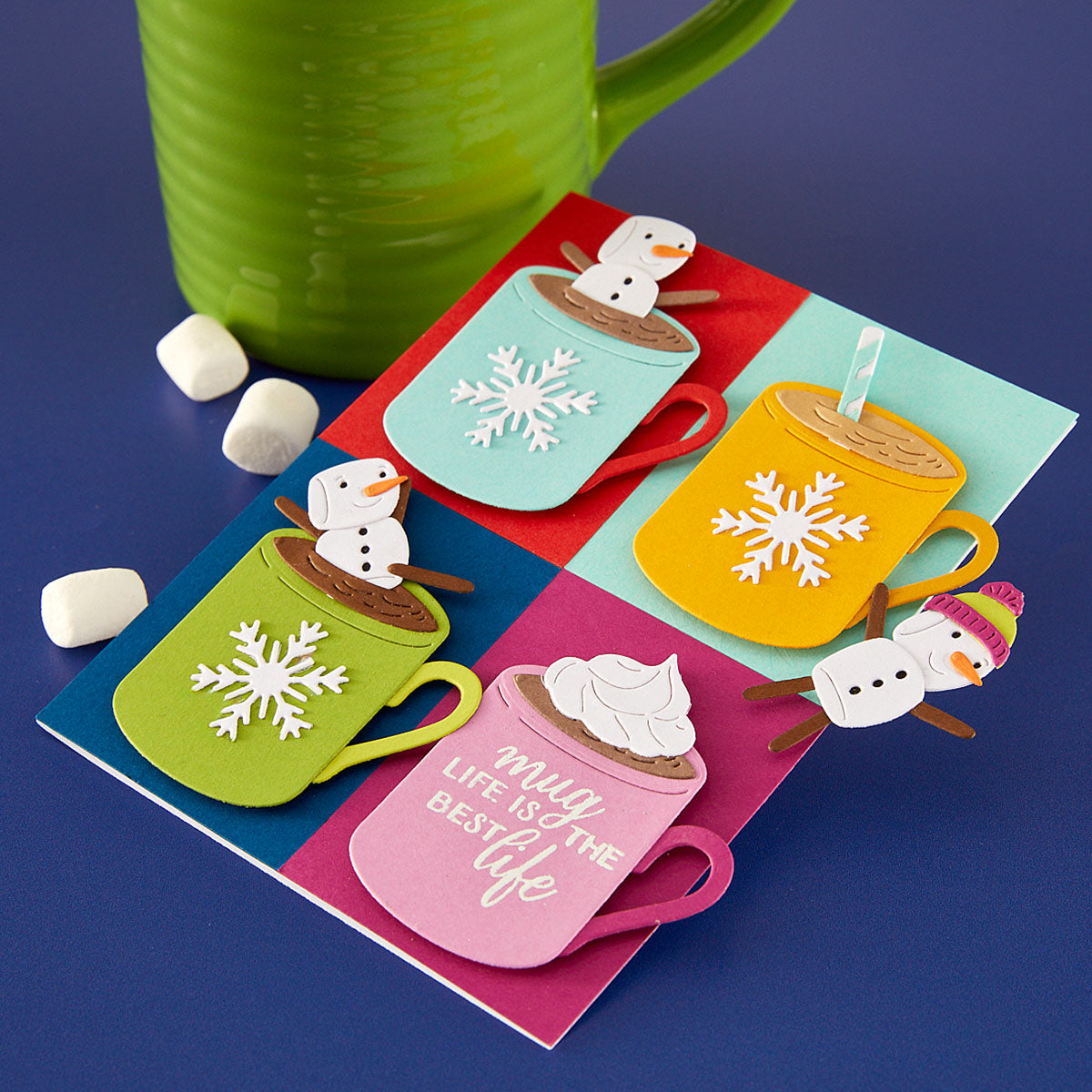 A colorful layered handmade card featuring four hot cocoa cups with snowflakes and snowmen, displayed on a blue surface with a green mug and marshmallows. Spellbinders Mugs and Kisses Sentiments Clear Stamp Set
