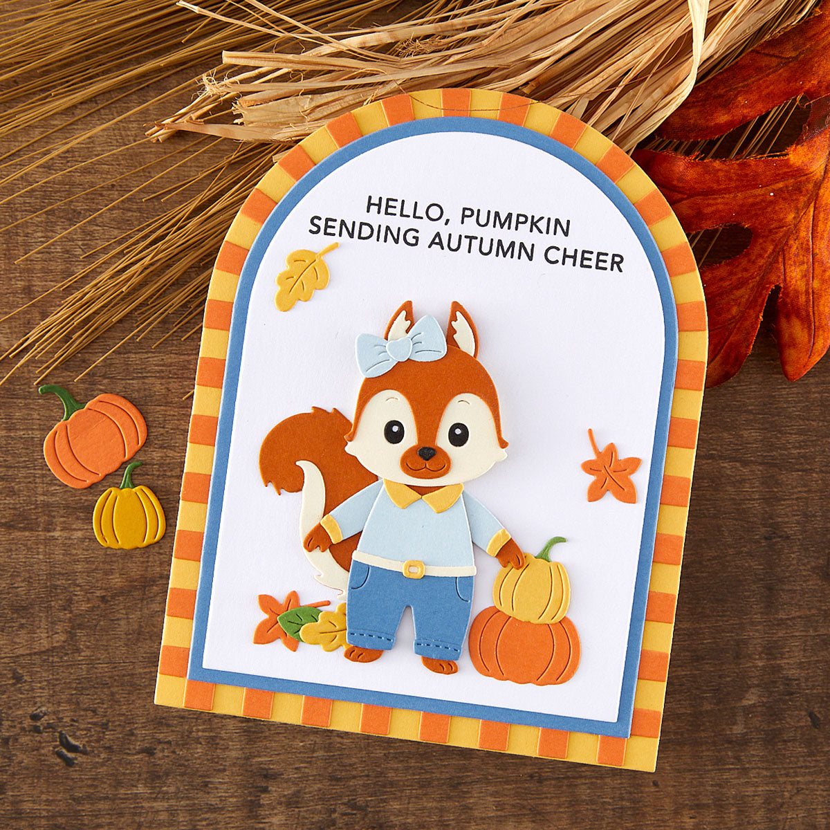 A handmade autumn-themed card featuring a layered paper fox character wearing blue pants and a light blue shirt, holding a small orange pumpkin. The card has a blue and orange striped border and reads 'HELLO, PUMPKIN SENDING AUTUMN CHEER'.