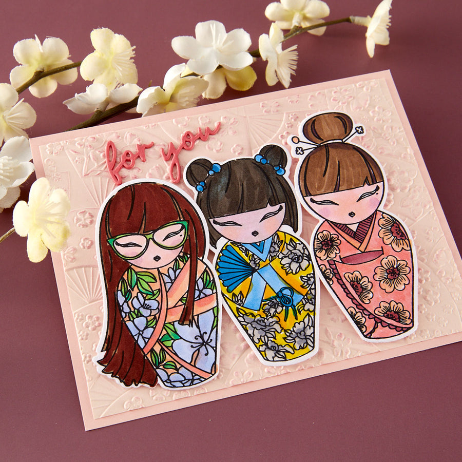 A decorative greeting card featuring three stylized illustrated girls in floral kimonos, placed on a mauve background with white flowers as props. The card has embossed fan motifs and the words 'for you' in pink script.