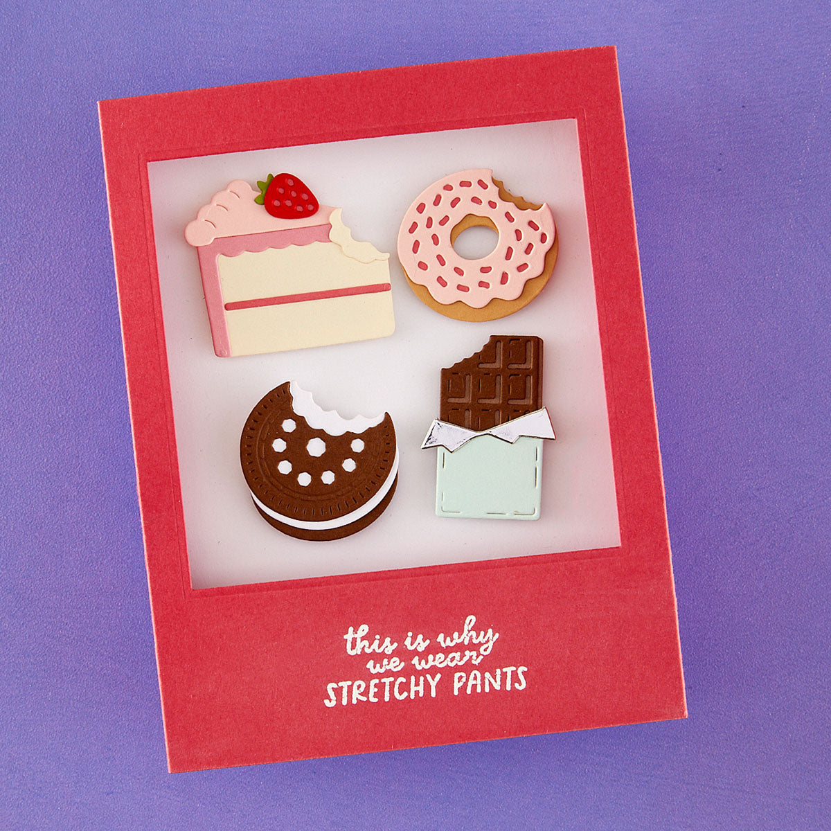 A handmade red greeting card with a clear window displaying four cute, 3D pastry illustrations: a slice of cake with a strawberry, a pink frosted donut, a chocolate cookie with a bite taken out, and a chocolate bar with its wrapper off. The bottom of the card features the white text 'this is why we wear STRETCHY PANTS'.