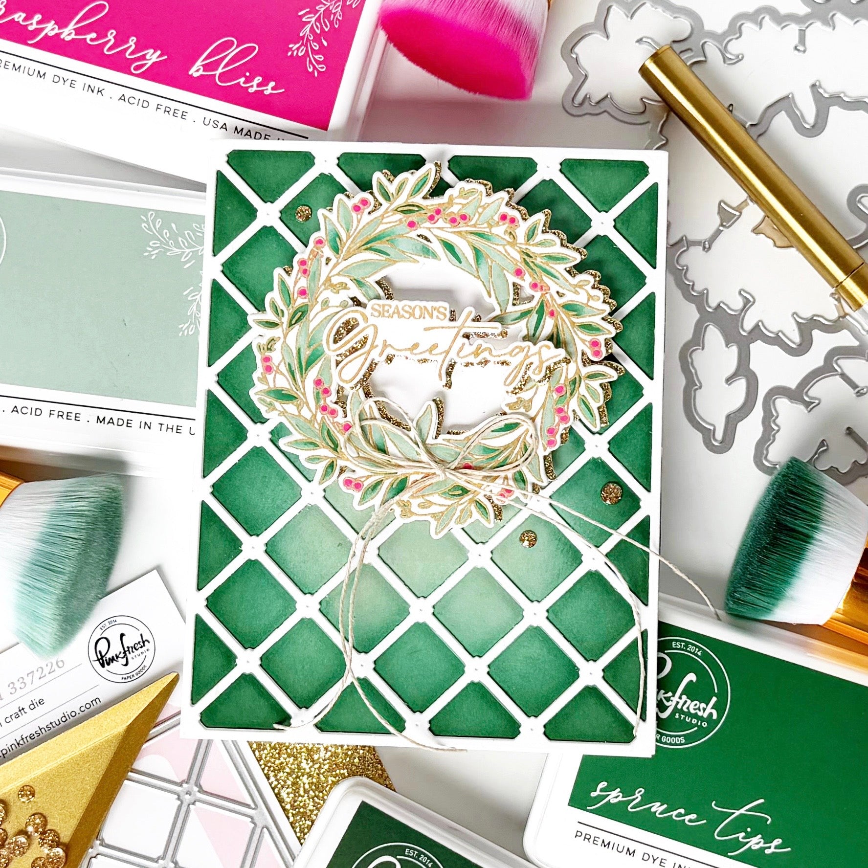 A handmade greeting card featuring a green and white lattice pattern with a layered wreath die-cut embellishment that reads 'Seasons Greetings.' The card is styled in a flat lay with crafting tools, brushes, and other card supplies around it.