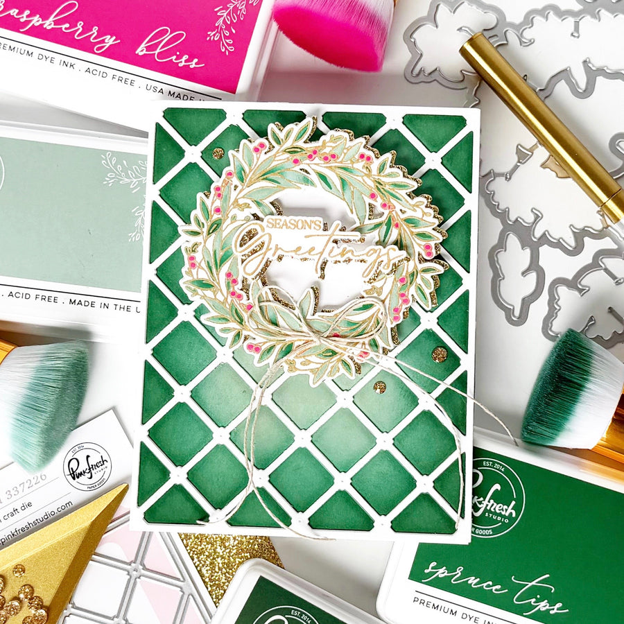 A handmade greeting card featuring a green and white lattice pattern with a layered wreath die-cut embellishment that reads 'Seasons Greetings.' The card is styled in a flat lay with crafting tools, brushes, and other card supplies around it.