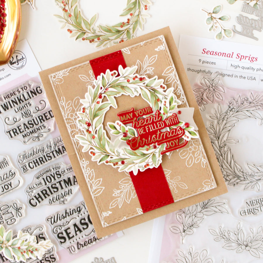 A handmade Christmas greeting card with a kraft paper base, a red ribbon, and a layered wreath embellishment with red berries, shown on a flat-lay surface alongside stamp sheets and floral stamp designs.