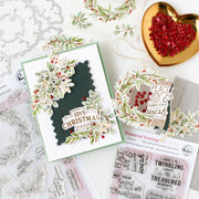 A styled flat-lay of handmade Christmas greeting cards and ornaments on a white background, surrounded by crafting tools and embellishments. The main product is a layered card with a dark green silhouette shape, white floral die-cut elements, and red berry accents, alongside a matching ornament and clear stamp sheets.