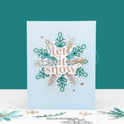 A handmade layered card featuring a die-cut snowflake design with the words 'Let it snow' in white script. The card has a light blue textured background, decorated with layered white, teal, and gold foil snowflakes, set against a solid teal background with a white surface below.
