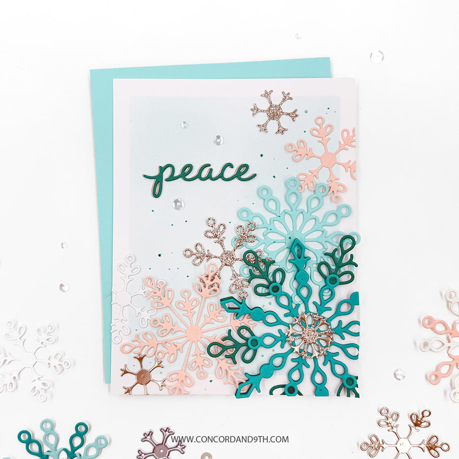 A handmade greeting card with layered die-cut snowflakes in teal, peach, and rose gold, featuring the word 'peace' in green script, shown with a light blue envelope on a white background with scattered decorative snowflakes.
