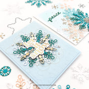 A handmade layered card featuring a large snowflake design with the words 'let it snow' in white, teal, and gold, displayed on a light blue card. A second card in the background shows a similar design with the word 'peace' in teal.