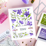 A handmade greeting card with purple flowers and green leaves on a white background, featuring the text 'Wishing you a beautiful day' on a banner. The card is styled in a flat-lay arrangement with crafting supplies, including Rhinestone Ink pads, thread, and stamps, on a light pink background.