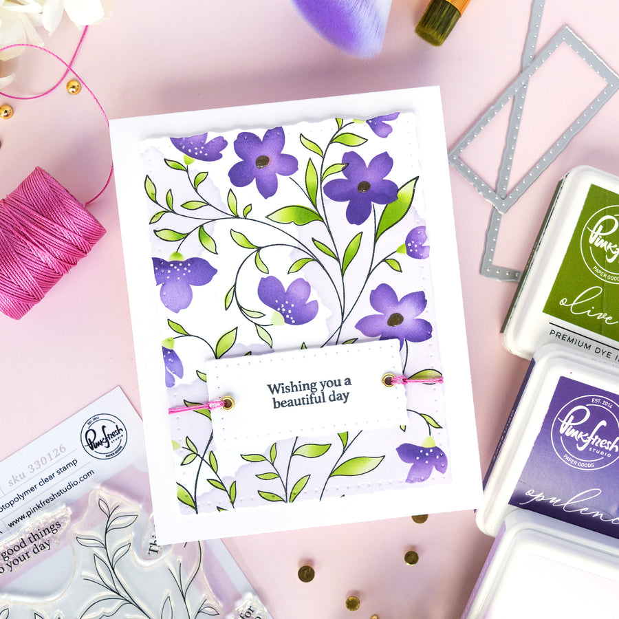 A handmade greeting card with purple flowers and green leaves on a white background, featuring the text 'Wishing you a beautiful day' on a banner. The card is styled in a flat-lay arrangement with crafting supplies, including Rhinestone Ink pads, thread, and stamps, on a light pink background.