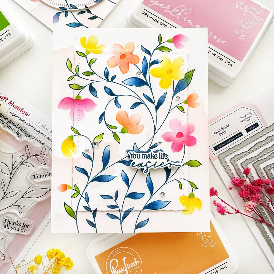 A handmade floral greeting card with blue leaves, yellow and pink flowers, and the phrase 'You make life easier' in the center, styled on a white surface with craft supplies including ink pads, stamps, and dried flowers.