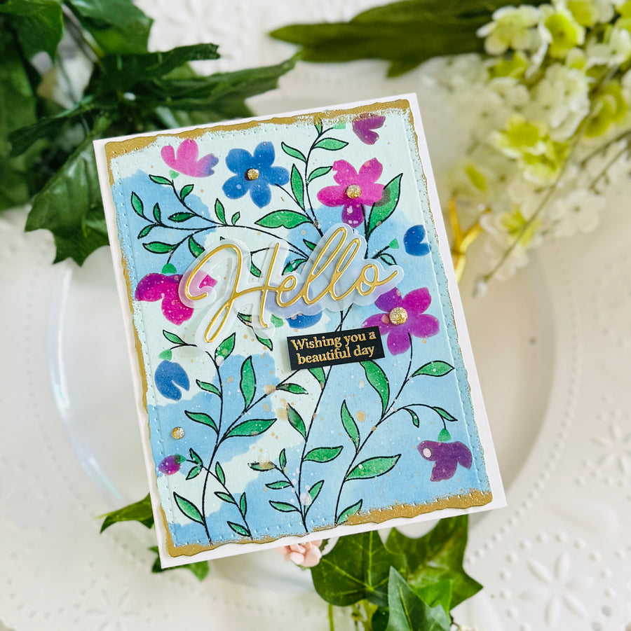 A handmade greeting card with a light blue background, decorated with hand-painted flowers in pink, purple, and blue, green leaves, gold foil script reading 'Hello', and a small black label that says 'Wishing you a beautiful day'. The card is styled on a white plate with green and white flowers.